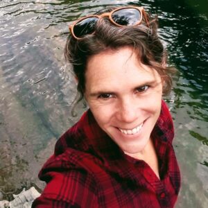 Amy Proctor - 5-8 Teacher | Oak Meadow. Amy is standing in front of a river. She's wearing a red flannel shirt and has brown sunglasses on her head.