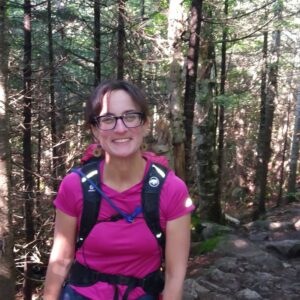 Alyssa Walker, an Oak Meadow teacher, is standing on a woody hiking trail smiling. She's wearing glasses, a bright pink hiking shirt, and a backpack