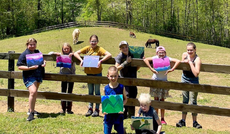 group of independent learning students showing off paintings next to horse pasture