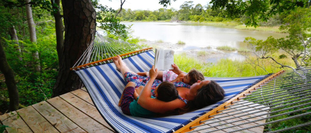 mom reading to her two kids while laying on a hammock by a river